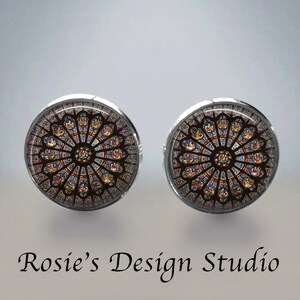 May include: A pair of silver earrings with a round glass cabochon featuring a stained glass window design. The window is a circular design with a central rosette and radiating spokes of colored glass.