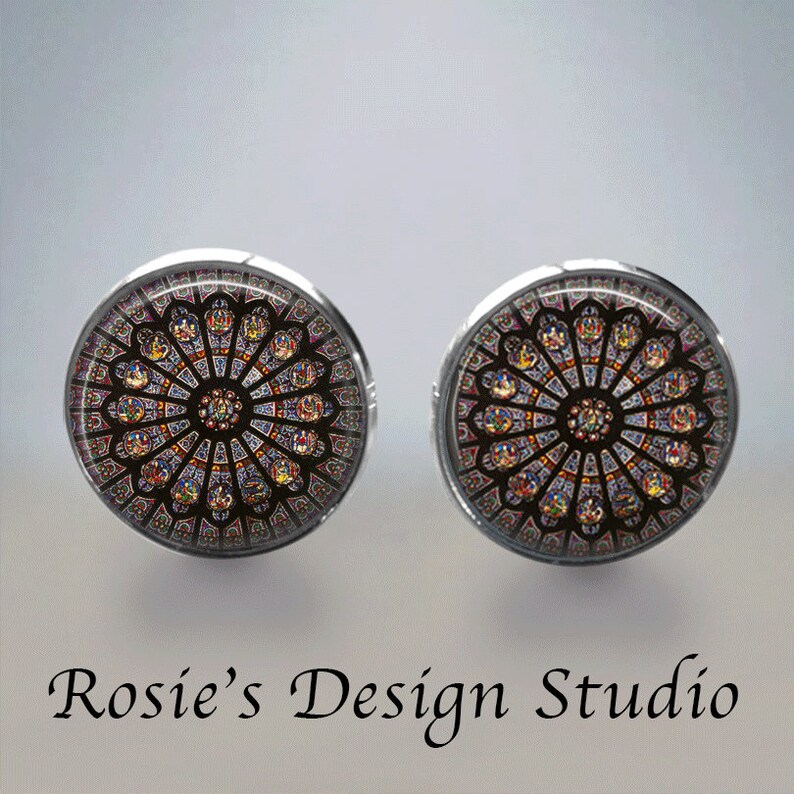 May include: A pair of silver earrings with a round glass cabochon featuring a stained glass window design. The window is a circular design with a central rosette and radiating spokes of colored glass.