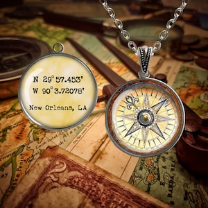 May include: A silver compass necklace with a vintage-style compass face. The compass face is cream-colored with black markings and a gold-colored needle. The back of the pendant has the coordinates "N 29°57.453' W 90°3.72078'" and the text "New Orleans, LA".