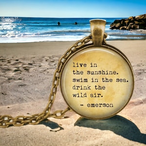 K&ouml;nnte beinhalten: Eine goldene Halskette mit einem runden Glasanh&auml;nger, auf dem ein Zitat von Ralph Waldo Emerson im Vintage-Stil steht: "live in the sunshine. swim in the sea. drink the wild air." Der Anh&auml;nger liegt auf einem Sandstrand mit dem Meer im Hintergrund.