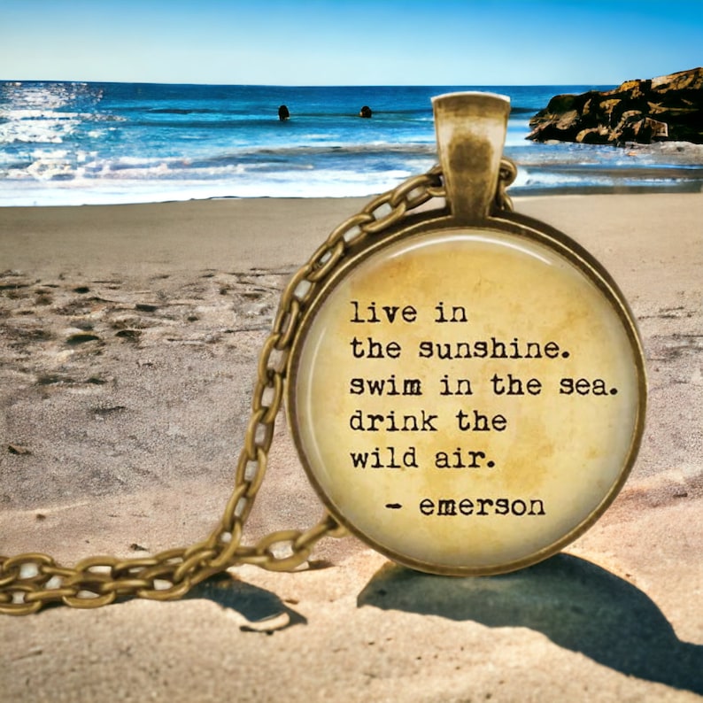 K&ouml;nnte beinhalten: Eine goldene Halskette mit einem runden Glasanh&auml;nger, auf dem ein Zitat von Ralph Waldo Emerson im Vintage-Stil steht: "live in the sunshine. swim in the sea. drink the wild air." Der Anh&auml;nger liegt auf einem Sandstrand mit dem Meer im Hintergrund.
