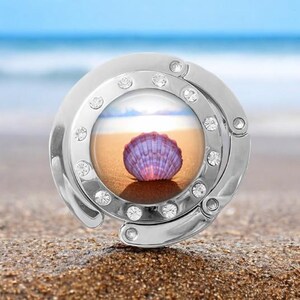 May include: A silver-toned, foldable purse hook with a clear round image of a seashell on a sandy beach. The hook is decorated with rhinestones.