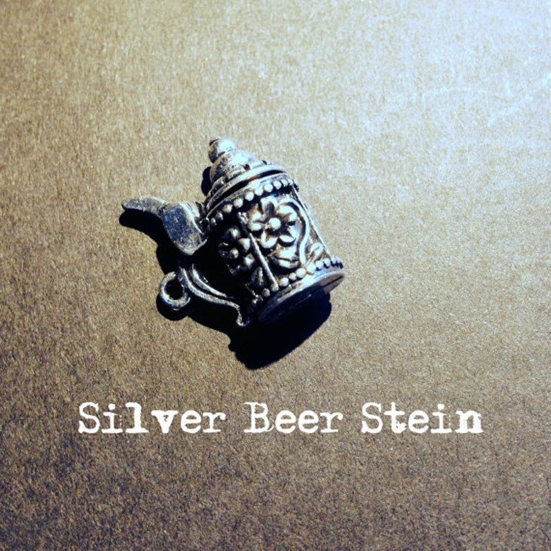 May include: A silver beer stein charm with intricate floral designs. The charm is a small, detailed replica of a traditional beer stein.