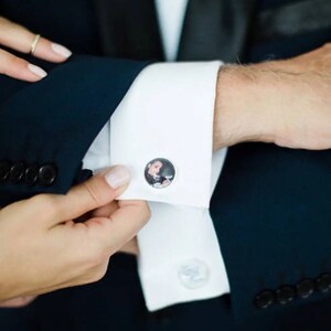 May include: A white dress shirt with a pair of cufflinks featuring a photo of a person. The cufflinks are on the left cuff of the shirt.