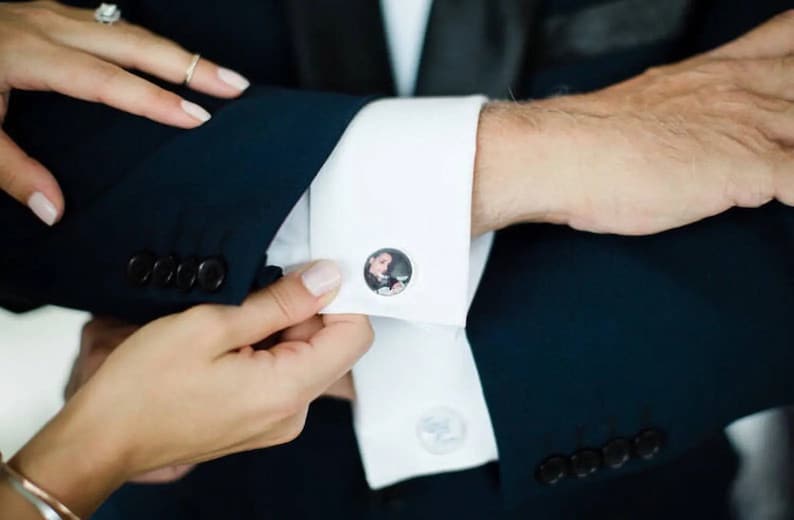 May include: A white dress shirt with a pair of cufflinks featuring a photo of a person. The cufflinks are on the left cuff of the shirt.