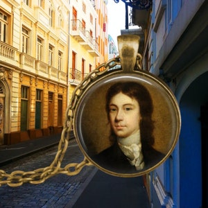 May include: A round, antique-style pendant necklace featuring a portrait of a man with long brown hair and a white cravat. The pendant is set in a gold-toned metal frame and hangs from a matching chain. The background shows a colorful street scene.