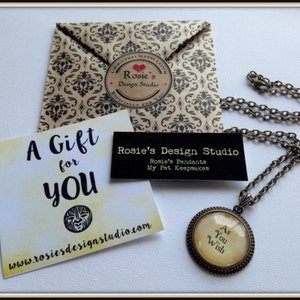 May include: A gold necklace with a round pendant that says "As You Wish". The pendant is on a brown background with a white card that says "A Gift for You" and a website address.