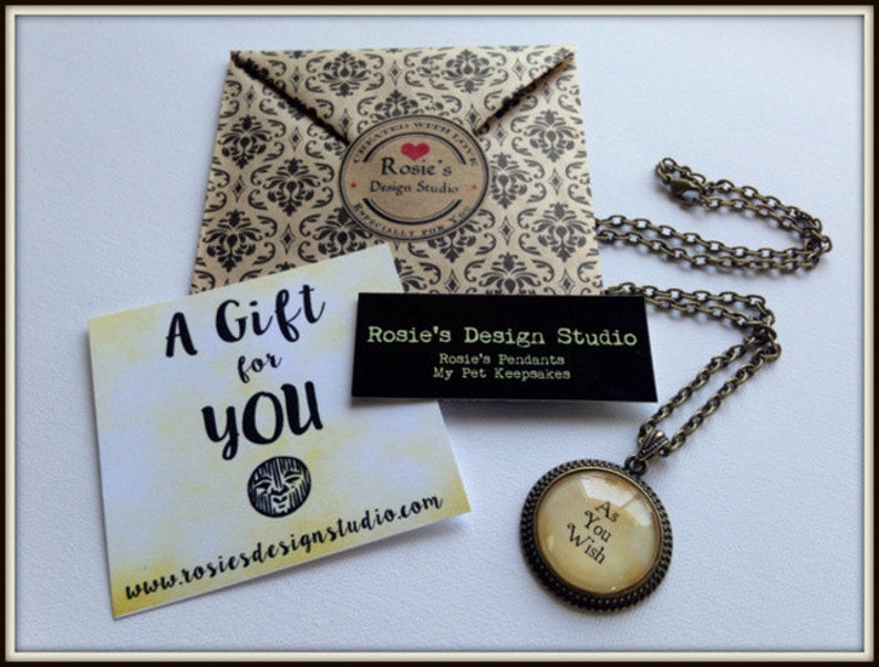 May include: A gold necklace with a round pendant that says "As You Wish". The pendant is on a brown background with a white card that says "A Gift for You" and a website address.