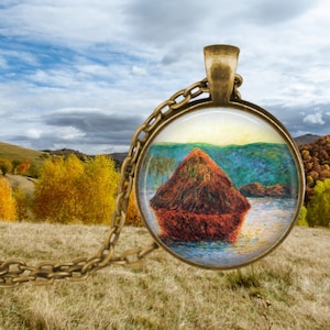 May include: A bronze chain necklace with a round glass pendant featuring a painting of a haystack in a field. The painting is in a style reminiscent of Impressionism, with visible brushstrokes and a focus on light and colour.