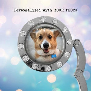 May include: A silver-toned, round, photo charm with a clear plastic window. The charm has a hinged clasp and is decorated with rhinestones. The window shows a photo of a smiling corgi dog wearing a blue collar with a bone-shaped tag.