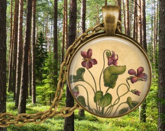 May include: A brass necklace with a round glass pendant featuring a vintage botanical illustration of purple and pink flowers with green leaves on a cream background.