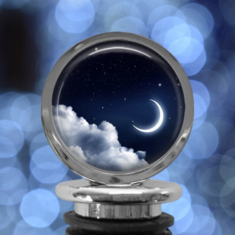 May include: A silver bottle stopper with a round, clear top featuring a night sky with a crescent moon and clouds. The background is a blue bokeh.