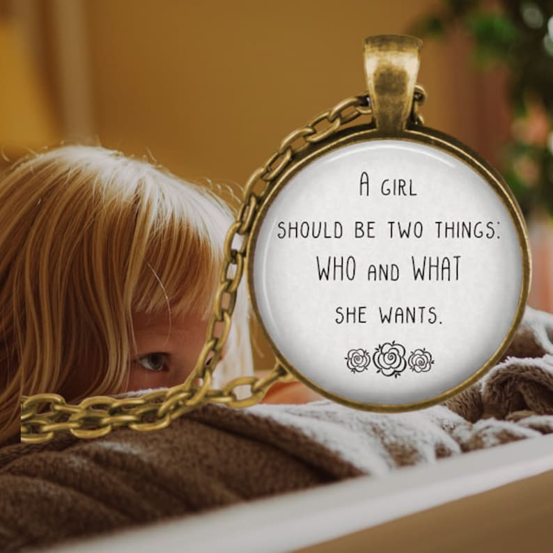 May include: A gold necklace with a round pendant that says "A girl should be two things: who and what she wants." The pendant is surrounded by three small rose designs.