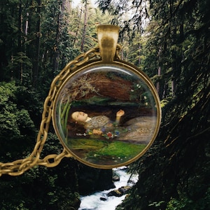 May include: A gold pendant necklace with a round glass cameo featuring a painting of a woman lying in a stream of water surrounded by flowers and greenery.
