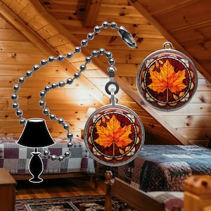 May include: A set of two decorative ceiling fan pull chains with a stained glass design featuring a fall leaf in shades of orange, yellow, and red. The chains are made of silver metal beads.