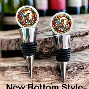 May include: Two silver-toned bottle stoppers featuring a circular top with a stained-glass style hummingbird design in red, green, and yellow. Each stopper has a tapered metal base with black rubber rings. The text New Bottom Style is visible.