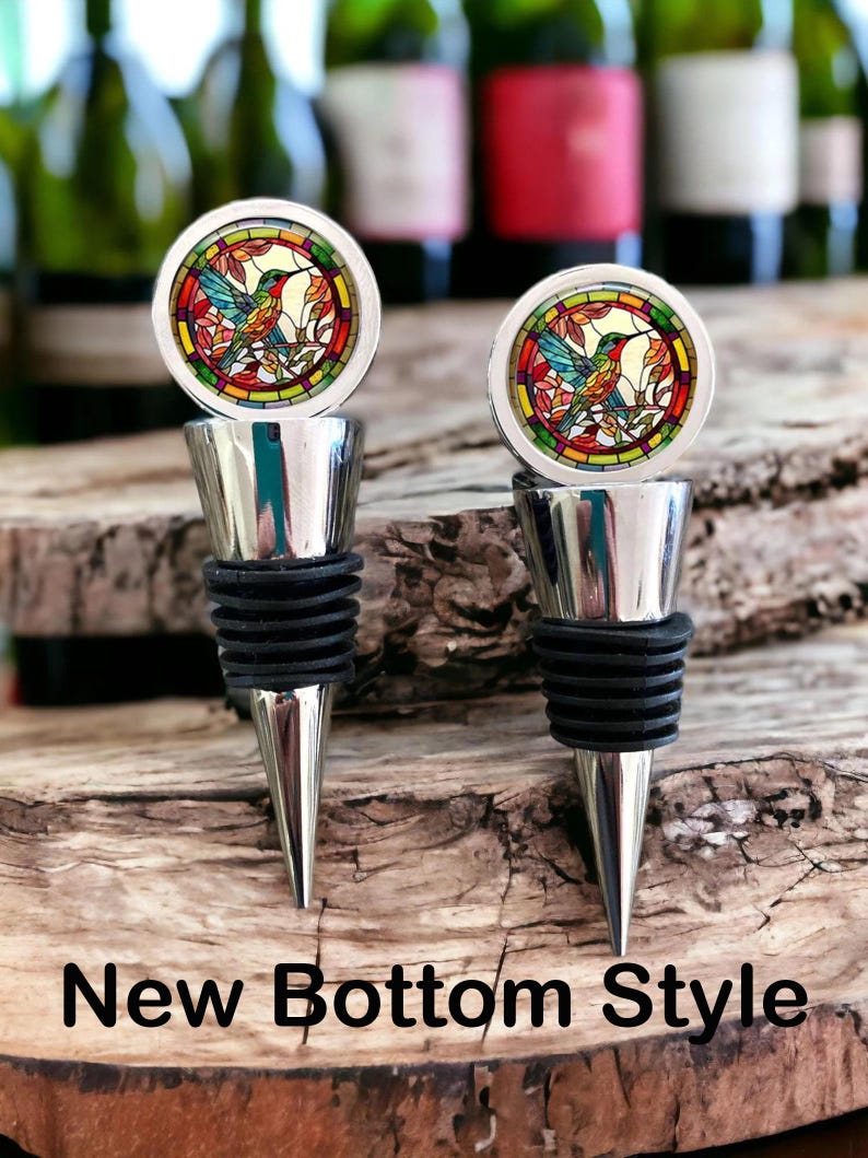 May include: Two silver-toned bottle stoppers featuring a circular top with a stained-glass style hummingbird design in red, green, and yellow. Each stopper has a tapered metal base with black rubber rings. The text New Bottom Style is visible.