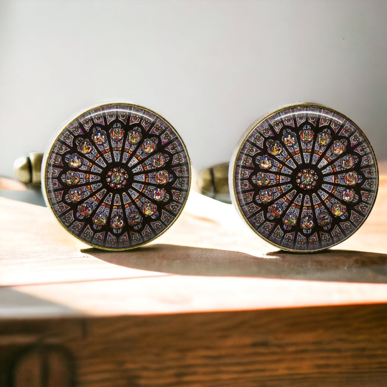 May include: A pair of cufflinks featuring a stained glass window design. The cufflinks are round and have a gold-tone finish. The stained glass window design is colorful and intricate, with shades of red, blue, yellow, and green.
