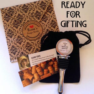 May include: A silver bottle stopper with a circular top featuring the text 'wine is bottled poetry' by Robert Louis Stevenson, a black drawstring pouch, a care instruction card with a vintage portrait, and a brown envelope with a damask pattern.
