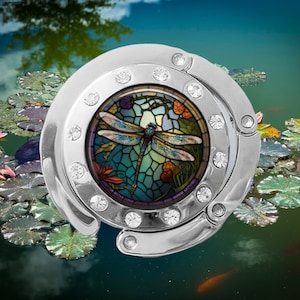 May include: A silver-toned metal purse hook with a round, clear crystal-encrusted frame. The frame features a stained glass design of a dragonfly with blue, green, and purple accents.