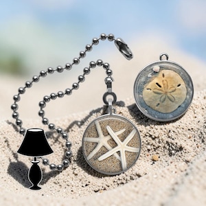 May include: Two decorative ceiling fan pull chains with beach-themed pendants. One pendant features two white starfish on sand, the other a sand dollar. Both are encased in clear resin with silver-tone metal settings. The chains are silver-coloured.