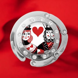 May include: A silver purse hook with a circular design featuring a Queen and King of Hearts playing card design. The hook is embellished with clear crystals and set against a red background.