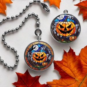 May include: Two silver and glass Halloween light pull chains with stained glass jack-o'-lantern designs. The chains are surrounded by autumn leaves.
