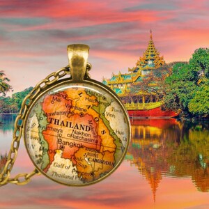 May include: A gold necklace with a round glass pendant featuring a vintage map of Thailand. The map shows major cities and geographical features, including the Mekong River and the Gulf of Thailand. The pendant is set against a backdrop of a traditional Asian temple reflected in a still body of water.