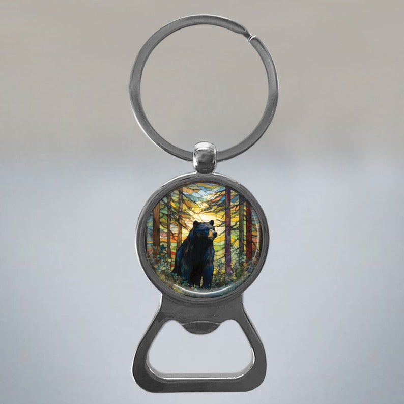 May include: A silver bottle opener keychain featuring a round, colourful image of a black bear standing in a forest. The image has a stained glass effect.