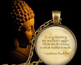 May include: A round bronze-toned pendant necklace featuring a cream-colored insert with the text: Every morning we are born again. What we do today is what matters most. ~ Gautama Buddha. The pendant is set against a dark background.