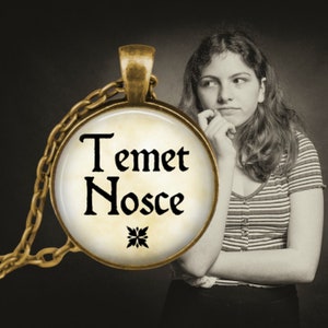 May include: A gold necklace with a round pendant featuring the words "Temet Nosce" in black lettering on a cream background. The pendant has a decorative design in the center.
