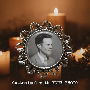 May include: A silver-toned photo frame with a black and white photo of a man inside. The frame is ornate and has a starburst design. The photo frame is sitting on a table with candles in the background.