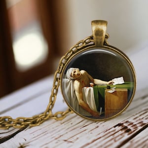 May include: A round glass pendant necklace with a bronze chain. The pendant features a painting of a man lying on a bed, with a quill pen in his hand, and a piece of paper on a table beside him. The painting is set against a dark background.