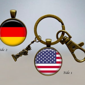 Op de afbeelding: Een bronzen sleutelhanger met een afneembare hanger met daarop het Vrijheidsbeeld. Aan de ene kant van de sleutelhanger zit een ronde glazen cabochon met de Amerikaanse vlag. Aan de andere kant zit een ronde glazen cabochon met de Duitse vlag. De tekst "Side 1" en "Side 2" staat op de afbeelding.
