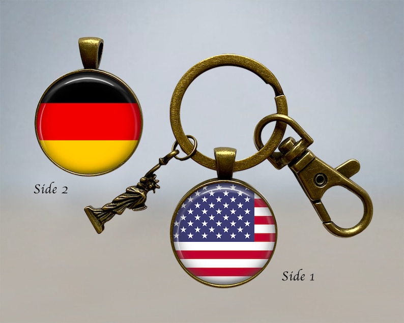 Op de afbeelding: Een bronzen sleutelhanger met een afneembare hanger met daarop het Vrijheidsbeeld. Aan de ene kant van de sleutelhanger zit een ronde glazen cabochon met de Amerikaanse vlag. Aan de andere kant zit een ronde glazen cabochon met de Duitse vlag. De tekst "Side 1" en "Side 2" staat op de afbeelding.