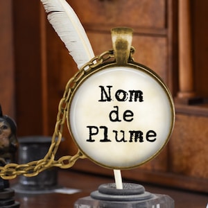 May include: A gold necklace with a round glass pendant featuring the text "Nom de Plume" in a vintage typewriter font on a cream background.