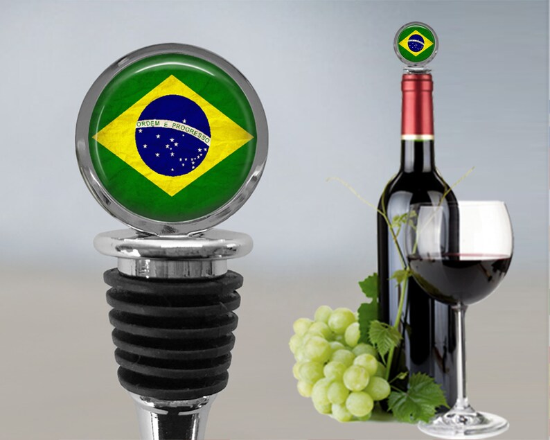 Op de afbeelding: Een zilveren flessenstop met een zwarte rubberen afdichting. De bovenkant van de stop heeft een cirkelvormig ontwerp met de Braziliaanse vlag. De vlag is groen, geel en blauw met een witte cirkel in het midden. De tekst "ORDEM E PROGRESSO" staat in wit op een blauwe band over het midden van de vlag.