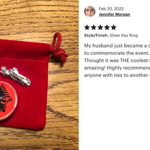 Op de afbeelding: Een zilveren sleutelhanger met een rode en zwarte Albanese vlag charme. De charme is bevestigd aan een zilveren sleutelhanger met een zilveren sleutelketting. De sleutelhanger zit in een rode fluwelen zakje.