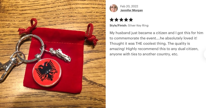 Op de afbeelding: Een zilveren sleutelhanger met een rode en zwarte Albanese vlag charme. De charme is bevestigd aan een zilveren sleutelhanger met een zilveren sleutelketting. De sleutelhanger zit in een rode fluwelen zakje.