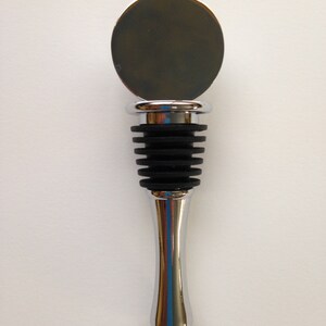 May include: A silver bottle stopper with a round, flat top and a black rubber stopper. The stopper is designed to fit into the neck of a wine bottle.