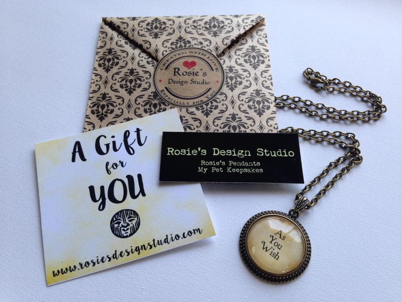 May include: A gift tag with the text "A Gift for You" and a website address. A brown paper envelope with a black and white pattern and a sticker that says "Rosie's Design Studio". A necklace with a round pendant that says "As You Wish".