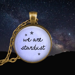 May include: A round pendant necklace with a bronze-colored chain. The pendant has a light blue background with the words "we are stardust" in black script, and three small star illustrations. The background is a starry night sky.