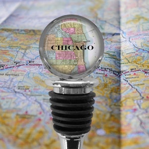 May include: A bottle stopper featuring a circular glass dome top with a vintage-style map of Chicago printed underneath. The stopper has a polished silver-tone metal base and a black, multi-tiered rubber gasket for sealing bottles securely.
