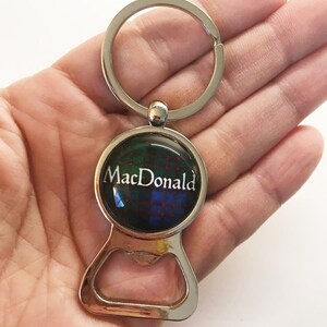 May include: Silver bottle opener keychain with a round, clear, glass charm featuring a tartan pattern and the text "MacDonald".