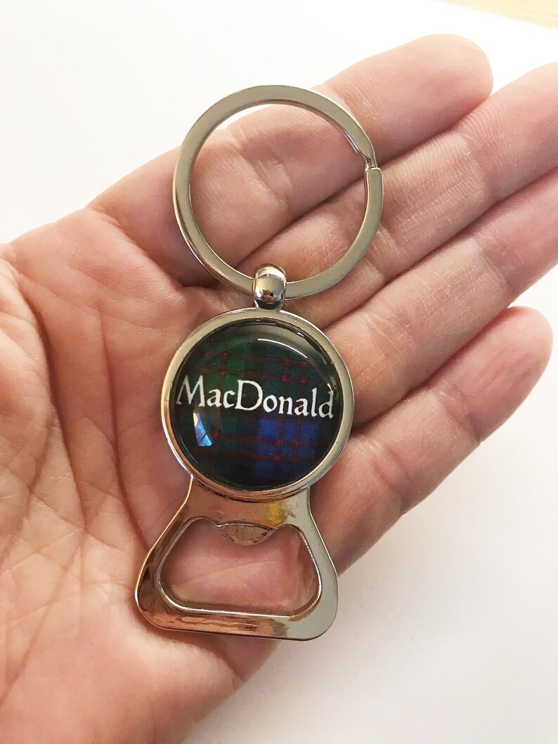 May include: Silver bottle opener keychain with a round, clear, glass charm featuring a tartan pattern and the text "MacDonald".