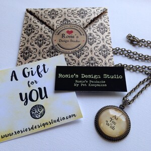 K&ouml;nnte beinhalten: Eine Geschenkkarte mit dem Text "A Gift for You" und einer Website-Adresse. Eine kleine schwarze Karte mit dem Text "Rosie's Design Studio" und dem Text "Rosie's Pendants My Pet Keepsakes". Eine goldene Halskette mit einem runden Anh&auml;nger, auf dem "As You Wish" steht.
