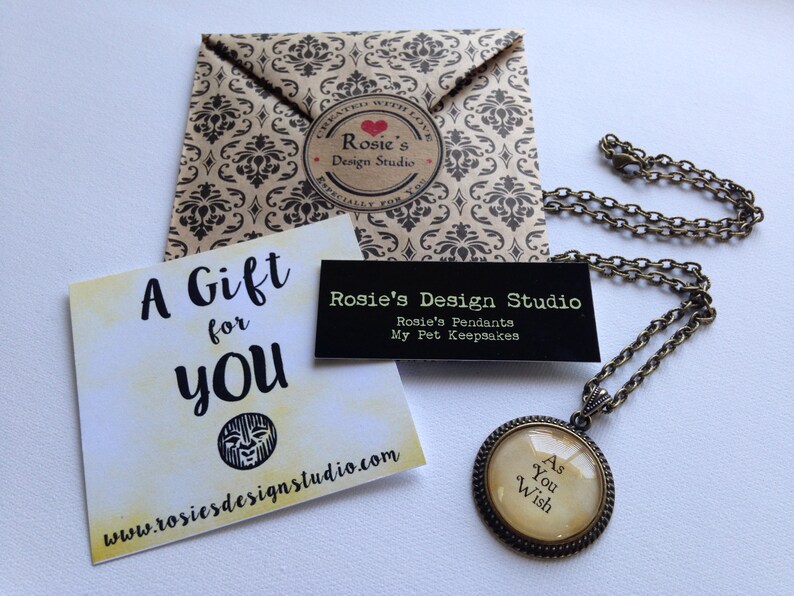 K&ouml;nnte beinhalten: Eine Geschenkkarte mit dem Text "A Gift for You" und einer Website-Adresse. Eine kleine schwarze Karte mit dem Text "Rosie's Design Studio" und dem Text "Rosie's Pendants My Pet Keepsakes". Eine goldene Halskette mit einem runden Anh&auml;nger, auf dem "As You Wish" steht.