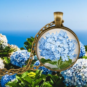 Peut inclure: Un collier avec un pendentif en forme de fleur d'hortensia bleue sur une chaîne en bronze. Le pendentif est un dôme de verre avec une illustration vintage d'une fleur d'hortensia bleue.