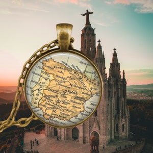 May include: A gold-toned pendant necklace featuring a circular charm with a vintage map of Spain. The pendant hangs from a chain. The background shows a church and a statue of Jesus Christ at sunset.