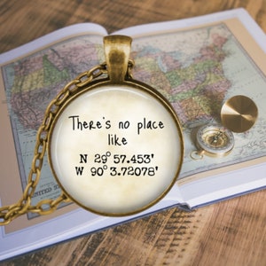 May include: A gold necklace with a round pendant featuring the text "There's no place like N 29° 57.453' W 90° 3.72078'" on a white background. The pendant is lying on a vintage map.